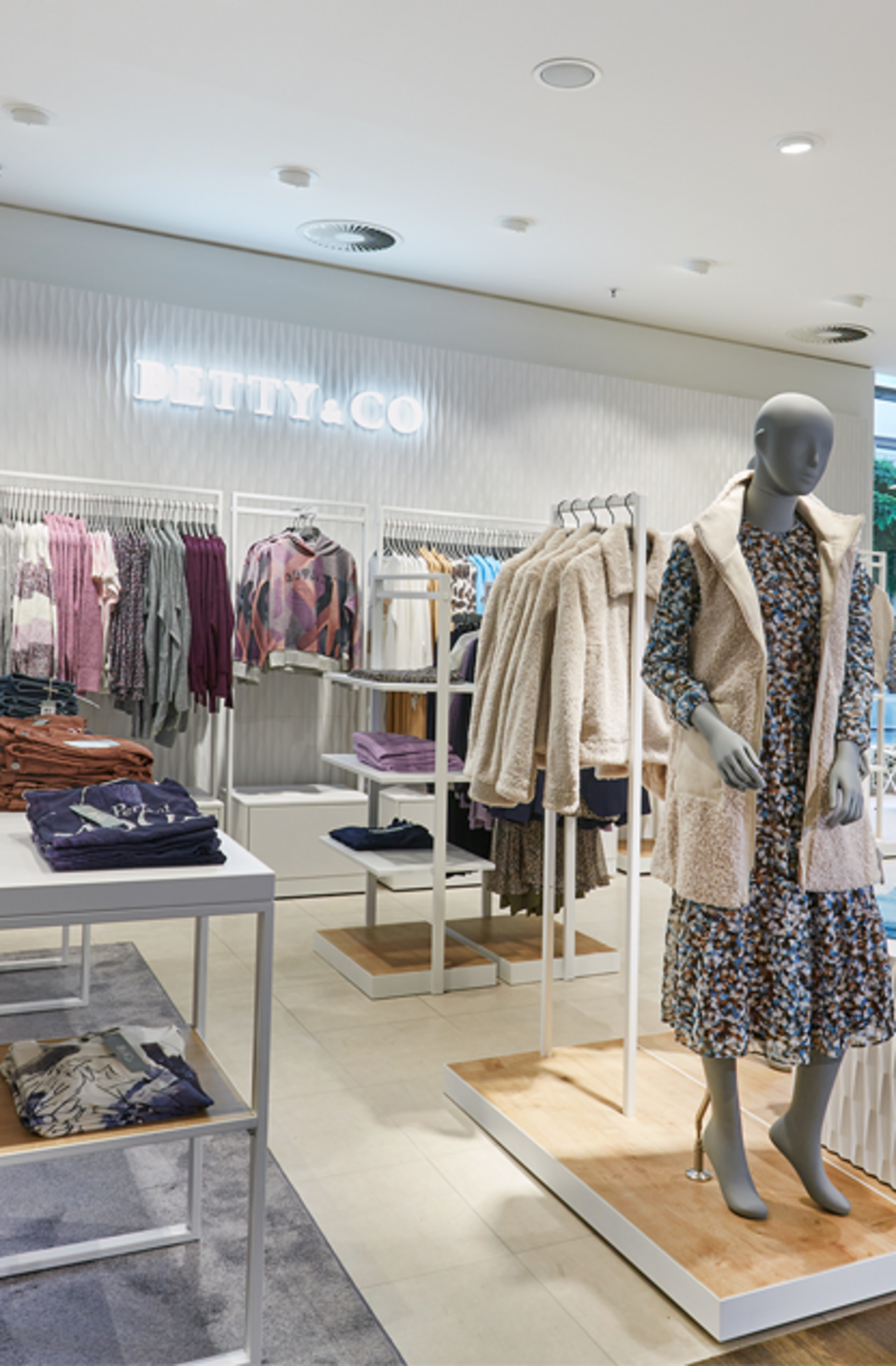 Innenansicht einer modernen Modeboutique der Marke BETTY & CO. Im Vordergrund zwei weiße Verkaufstische mit ordentlich gefalteter Damenbekleidung. Im Hintergrund Kleiderstangen mit Blusen, Jacken und Pullovern. Rechts stehen zwei Schaufensterpuppen mit herbstlicher Kleidung auf Podesten.