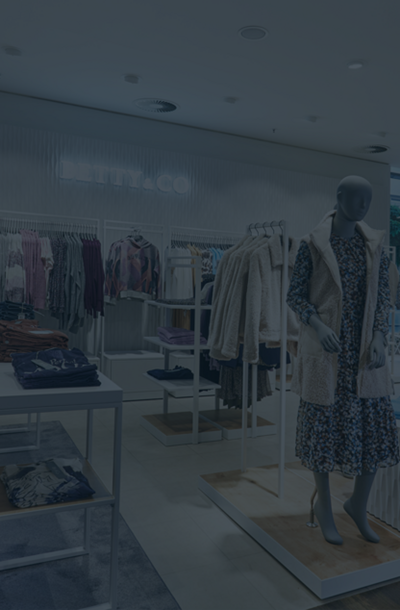 Mouse-Over: Innenansicht einer modernen Modeboutique der Marke BETTY & CO. Im Vordergrund zwei weiße Verkaufstische mit ordentlich gefalteter Damenbekleidung. Im Hintergrund Kleiderstangen mit Blusen, Jacken und Pullovern. Rechts stehen zwei Schaufensterpuppen mit herbstlicher Kleidung auf Podesten.